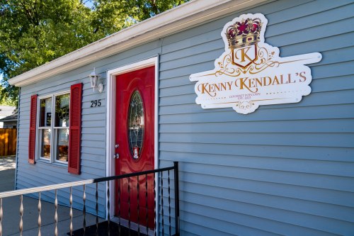 Exterior of a blue house with a red door and a sign reading "Kenny Kendall's" featuring a crown logo.