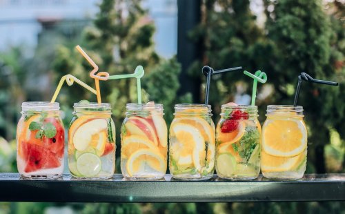 Five jars filled with colorful fruit-infused drinks, each with a straw, set against a green backdrop.