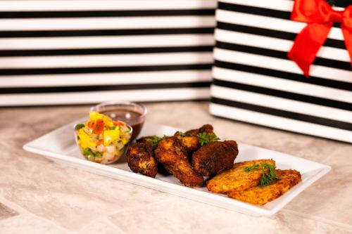 A plate of crispy chicken wings, plantain slices, and a fresh salsa, with a dipping sauce, set against striped gift boxes.