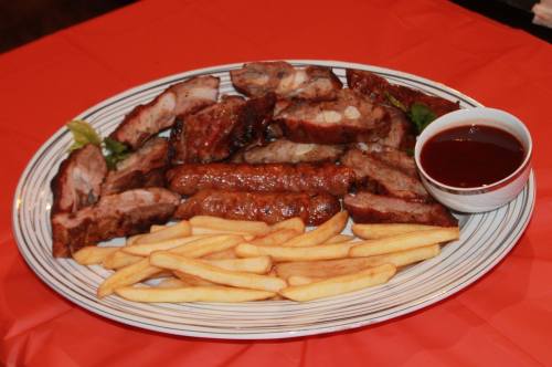 A platter of grilled meats including ribs and sausage, served with French fries and a small bowl of barbecue sauce.