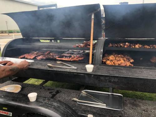 A large barbecue grill with smoke, featuring various meats cooking on multiple racks. Tongs and plates are nearby.