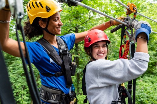 Two women in helmets prepare for ziplining, smiling and enjoying the outdoor adventure in a lush green setting.