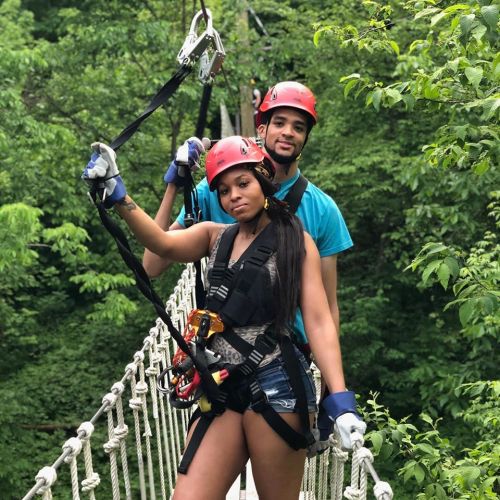 Two people in helmets and harnesses stand on a zip line platform surrounded by lush green trees.