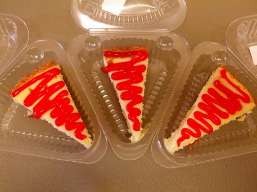 Three slices of cheesecake in clear plastic containers, topped with red sauce, arranged on a gray surface.