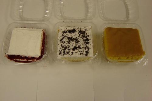 Three dessert squares in clear containers: red velvet, cookies and cream, and lemon cake.