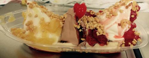 A plastic dish with three scoops of ice cream topped with nuts, fruit, and a cherry. Flavors include chocolate and strawberry.