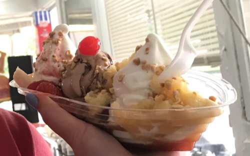 A hand holding a colorful ice cream sundae with three scoops, whipped cream, nuts, and a cherry on top.