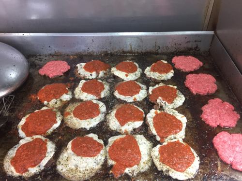 Cooking patties on a grill, some topped with red sauce, alongside raw ground meat in a commercial kitchen setting.
