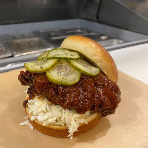 A crispy fried chicken sandwich topped with pickles and coleslaw on a soft bun, served on a paper wrapper.