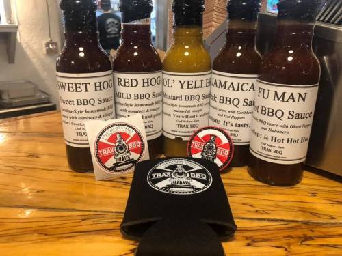 Five bottles of BBQ sauce on a wooden table, with stickers and a koozie from Trax BBQ.