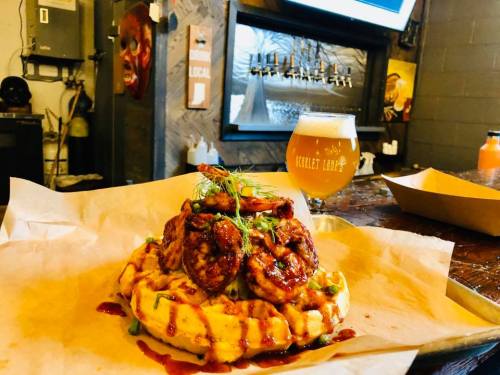 A plate of shrimp and waffles drizzled with sauce, accompanied by a glass of beer in a cozy bar setting.