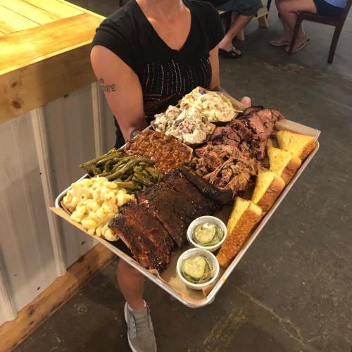 A person holds a large tray of barbecue, featuring ribs, pulled pork, sides, and bread.