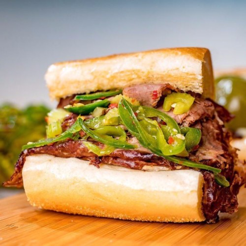 A close-up of a sandwich filled with sliced beef, jalapeños, and pickles on a soft bun, served on a wooden board.