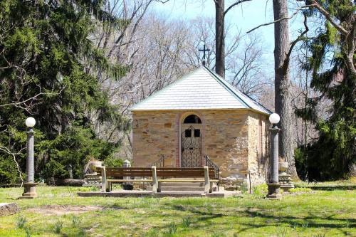 A small stone chapel with a sloped roof, surrounded by trees and a grassy area, featuring a bench in front.