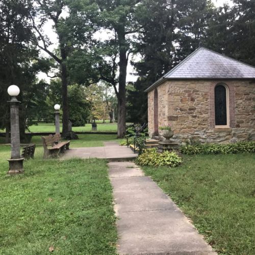 A stone building with a gray roof surrounded by trees, with a pathway leading to it and benches nearby.