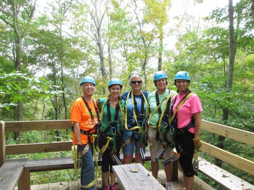 Five women in harnesses and helmets stand on a wooden platform in a forest, smiling and ready for an adventure.