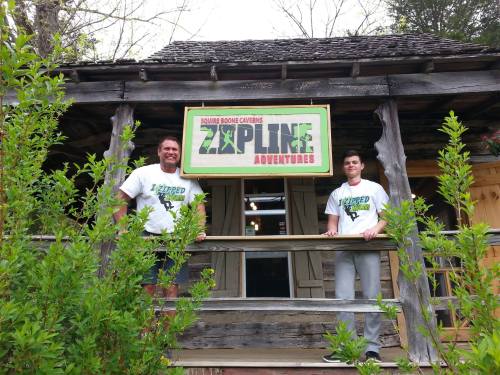 Two people stand on a porch in front of a wooden building with a sign that reads "Zipline Adventures."