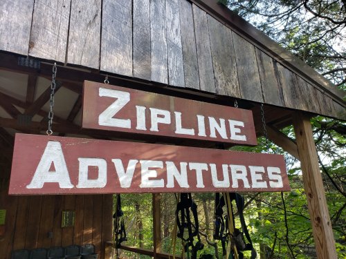 Sign for "Zipline Adventures" hanging from a wooden structure surrounded by trees.