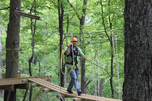 A person in a helmet and harness walks on a rope bridge among green trees in a forested area.