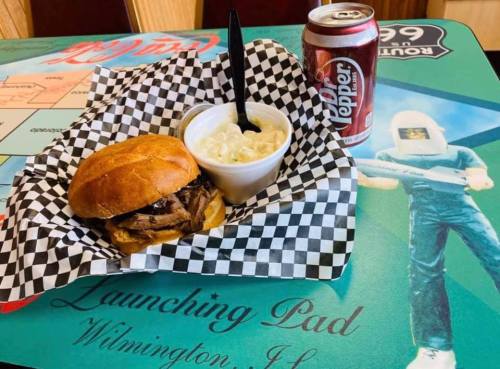 A sandwich with pulled meat on a checkered plate, a side of macaroni salad, and a can of Dr Pepper on a colorful table.