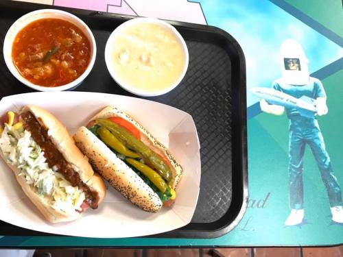 A tray with two hot dogs topped with condiments, a bowl of chili, and a bowl of soup on a colorful table.