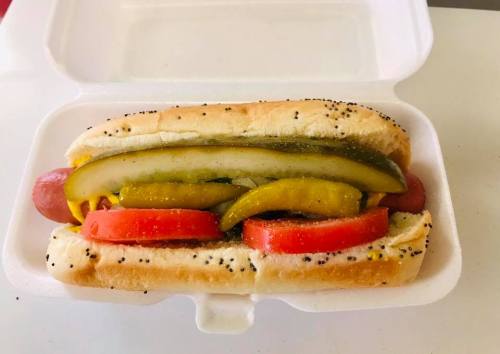 A hot dog in a bun topped with mustard, pickles, and tomato slices, served in a foam container.