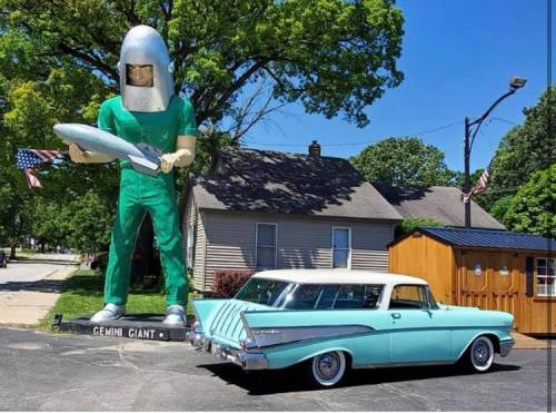 A large statue of a giant in a helmet holding a rocket stands next to a vintage blue car in a small town setting.