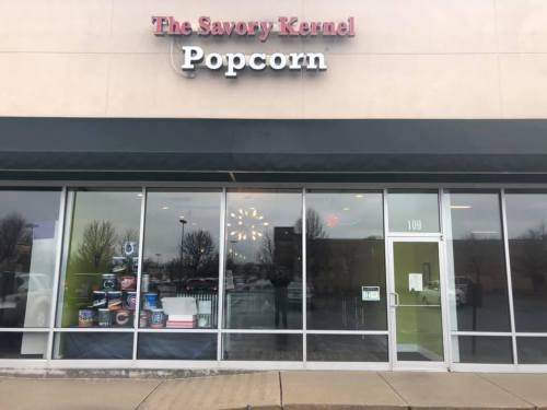 Storefront of The Savory Kernel Popcorn, featuring large windows and a variety of popcorn containers displayed inside.