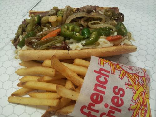 A loaded sandwich with beef, peppers, and onions, served with a side of crispy French fries.