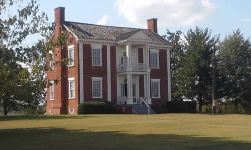 A historic two-story brick house with a porch, surrounded by green grass and trees under a blue sky.