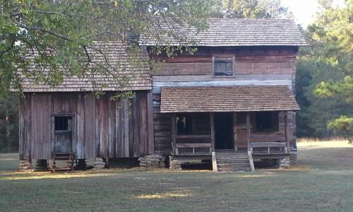 A rustic wooden house with a sloped roof, featuring a porch and surrounded by trees in a grassy area.