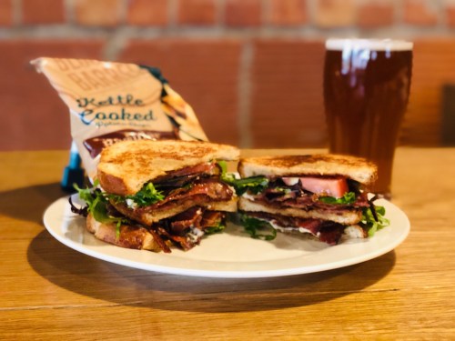 A hearty sandwich with layers of meat and greens, served with kettle-cooked chips and a glass of beer on a wooden table.