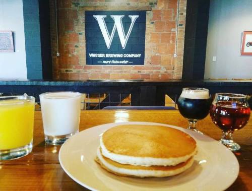 A plate of pancakes with three small glasses of drinks in front of a brewery logo on a brick wall.
