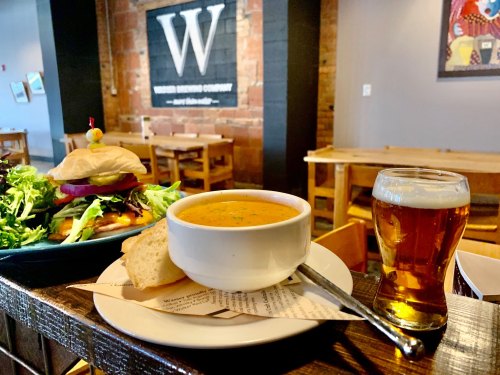 A bowl of soup with a side of bread, a burger, salad, and a glass of beer on a table in a cozy restaurant.