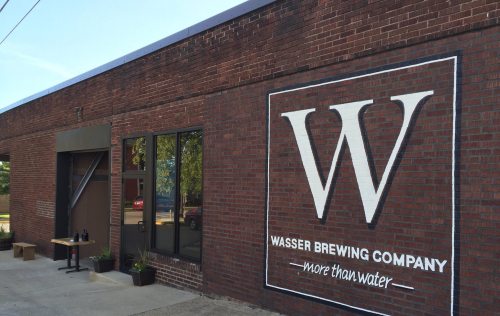 Exterior of Wasser Brewing Company featuring a large logo on a brick wall and a small table outside.