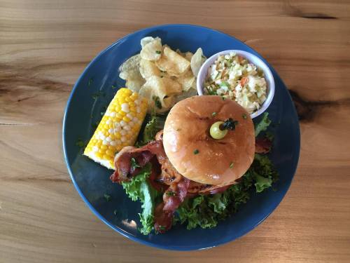 A plate with a bacon burger, corn on the cob, coleslaw, and potato chips on a wooden table.