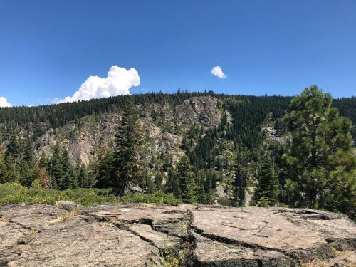 A rocky outcrop overlooks a lush green forested mountain under a clear blue sky with fluffy white clouds.