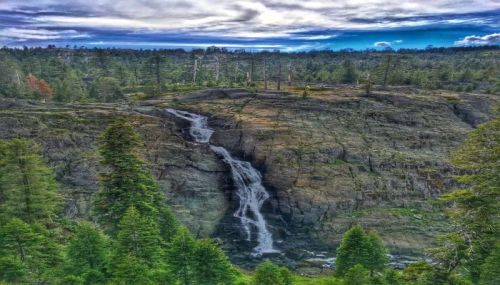 A scenic view of a waterfall cascading over rocky terrain, surrounded by lush greenery and a cloudy sky.