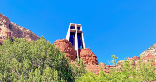 A modern building stands atop red rock formations against a clear blue sky, surrounded by greenery.