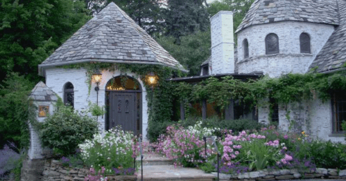 Charming cottage with a stone roof, surrounded by vibrant flowers and lush greenery. Warm lights illuminate the entrance.