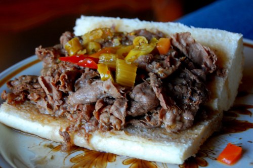 A plate with a sandwich filled with shredded beef, topped with peppers and onions on soft white bread.