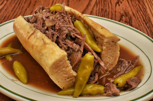 A hearty sandwich filled with shredded beef, served with yellow peppers and a side of savory broth.