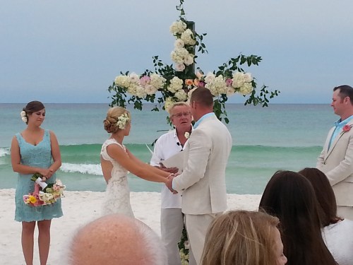 A beach wedding ceremony with a couple exchanging vows in front of a floral cross, surrounded by guests and attendants.