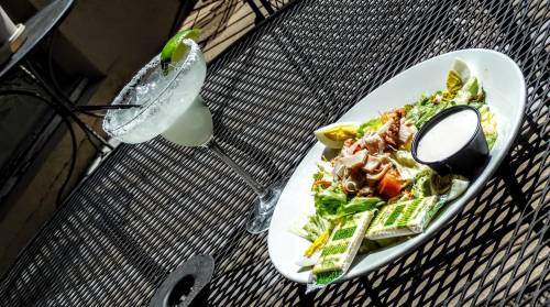 A plate of salad with grilled chicken and a side of ranch dressing, next to a margarita on a black metal table.