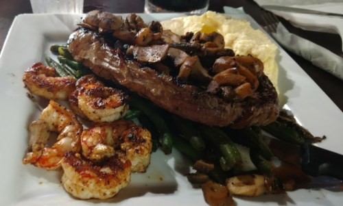 A plate featuring a grilled steak topped with mushrooms, shrimp, green beans, and creamy mashed potatoes.