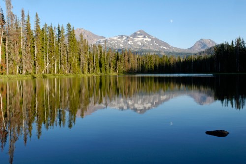 A serene lake reflects mountains and trees under a clear blue sky, with a hint of moon visible.