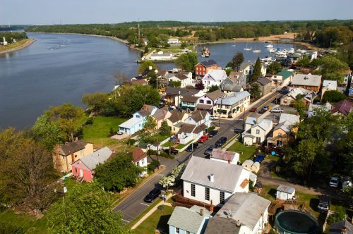 Aerial view of a riverside town with colorful houses, a road, and boats on the water. Lush greenery surrounds the area.