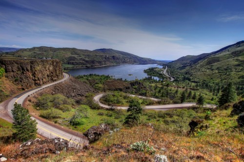 A winding road curves along a river, surrounded by lush green hills and a clear blue sky.