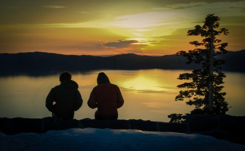Two people sit on a ledge, silhouetted against a sunset over a calm lake, with mountains in the background.