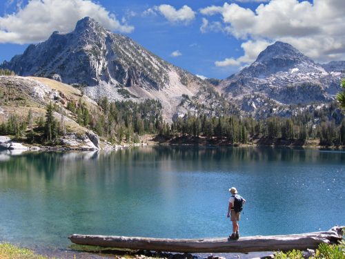 A hiker stands on a log by a serene lake, surrounded by mountains and lush trees under a blue sky with clouds.